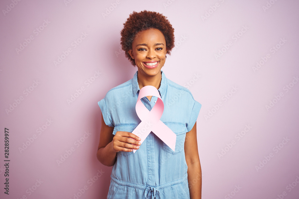 Young african american woman holding brest cancer ribbon over isolated pink background with a happy face standing and smiling with a confident smile showing teeth